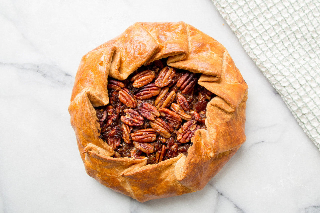 A top-down view of a rustic open faced pecan pie with golden-brown flaky pastry crust and glossy browned pecans, presented on a white marble surface with a textured white cloth in the corn